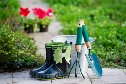 A close-up view of a garden pathway in a residential outdoor space, featuring a pair of dark rubber gardening boots with a decorative trim, hanging on a small hook. Nearby, a white plastic bucket with a metal handle holds gardening tools with wooden handles, including a trowel and a hand rake. Behind the bucket, two flower pots contain vibrant pink and red petunias, adding a splash of colour against the green foliage. The pathway is paved with rectangular stones, with the surrounding garden bed filled with lush, green low-growing plants and shrubs. In the background, a slightly blurred lawn area can be seen, bordered by hedge-like plants, suggesting a well-maintained garden space suitable for general gardening or lawn care services in the Earl's Court area. The scene appears to be in good weather, with natural daylight illuminating the scene, highlighting the textures of the garden elements and tools, providing a natural and inviting outdoor environment for gardening work.