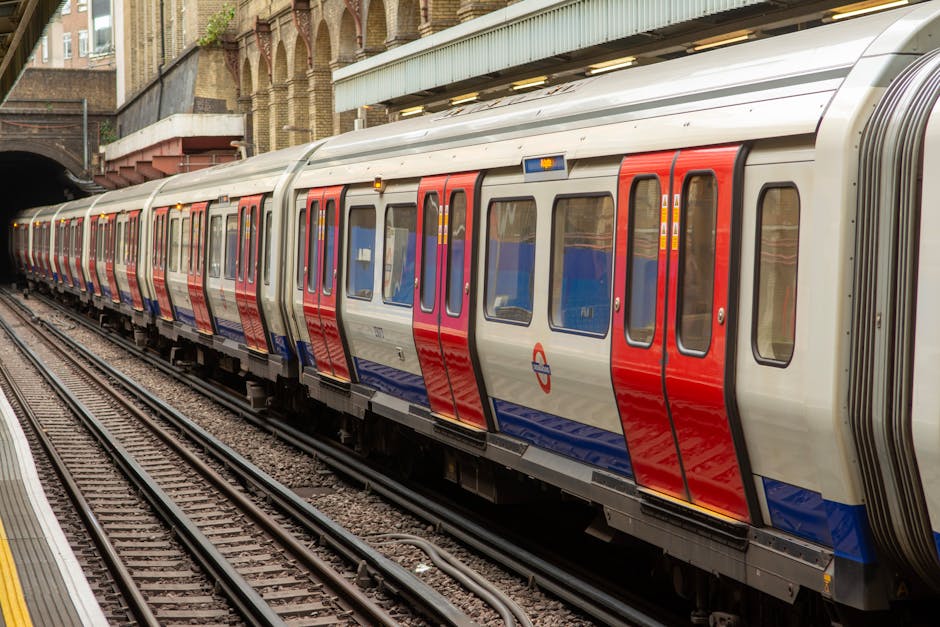 A modern London Underground train with a silver body and distinctive red, blue, and purple panel accents is stationed on the platform near Earls Court. The train's windows reflect the surrounding envi