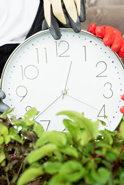 Close-up of a gardener's gloved hands holding a round, white wall clock with black numerals and hands, partially obscured by lush green garden plants and foliage in the foreground. The garden features